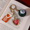 fidget keychain with gold heart photo frame, enamel orange juice charm, and 8-ball de-stress ball on a book