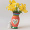pink folk floral vase with a white cat 'tipping' it over with yellow daffodils