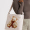 model carrying tan horizontal zip bag with teddy bear with a red bow around the neck