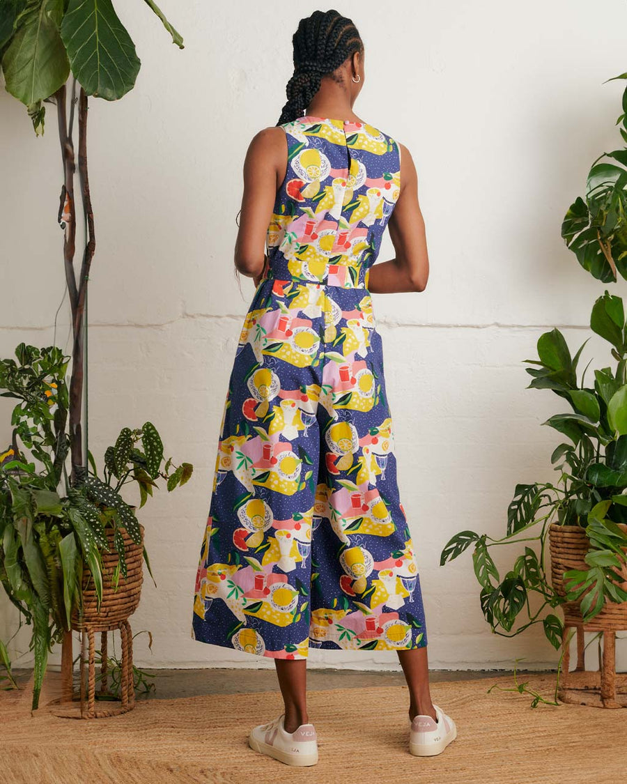 back view of model wearing blue jumpsuit with picnic food print and tie waist