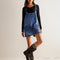 model wearing blue denim overall dress with front patch pockets and unique strap closure