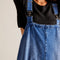 up close of model wearing blue denim overall dress with front patch pockets and unique strap closure
