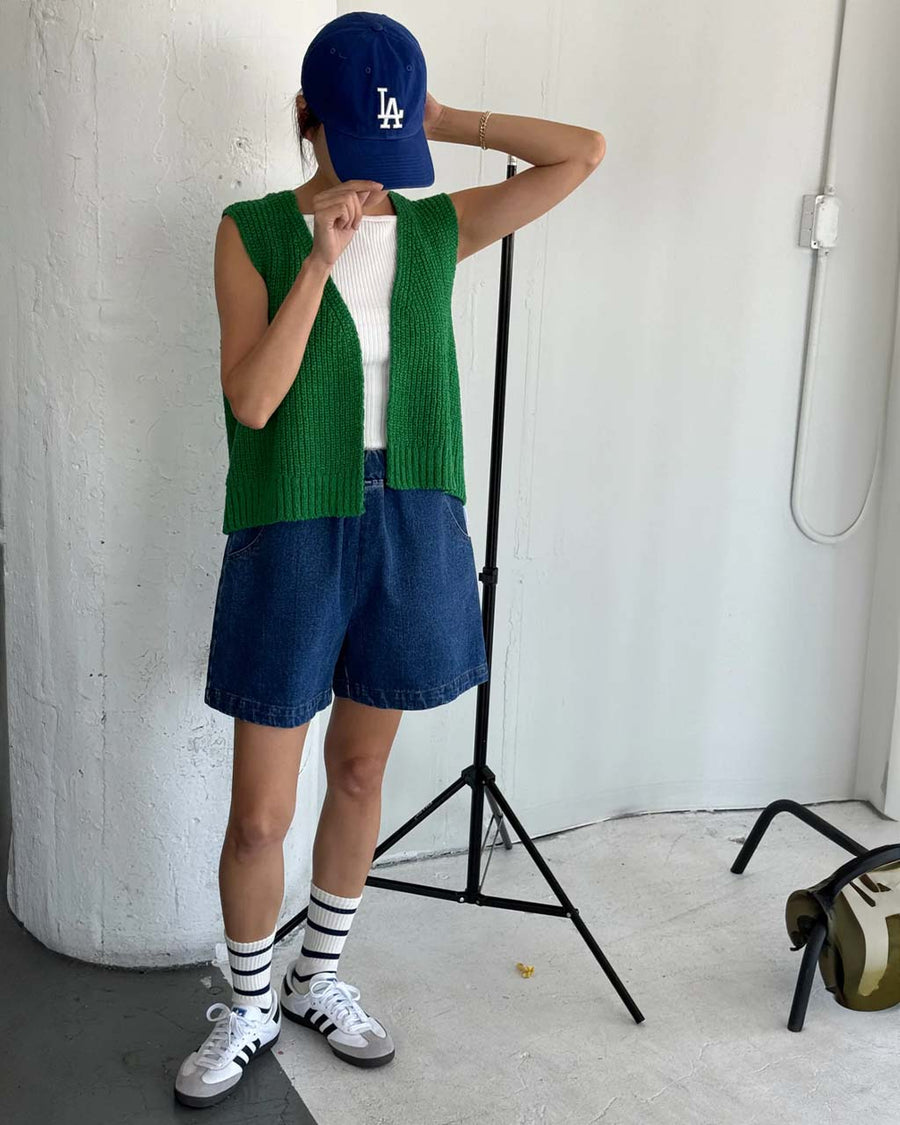 model wearing denim shorts with pockets and elastic waist with green knit vest, white top, and baseball cap