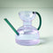 lavender abstract shaped watering can with teal colored spout and handle on a green background