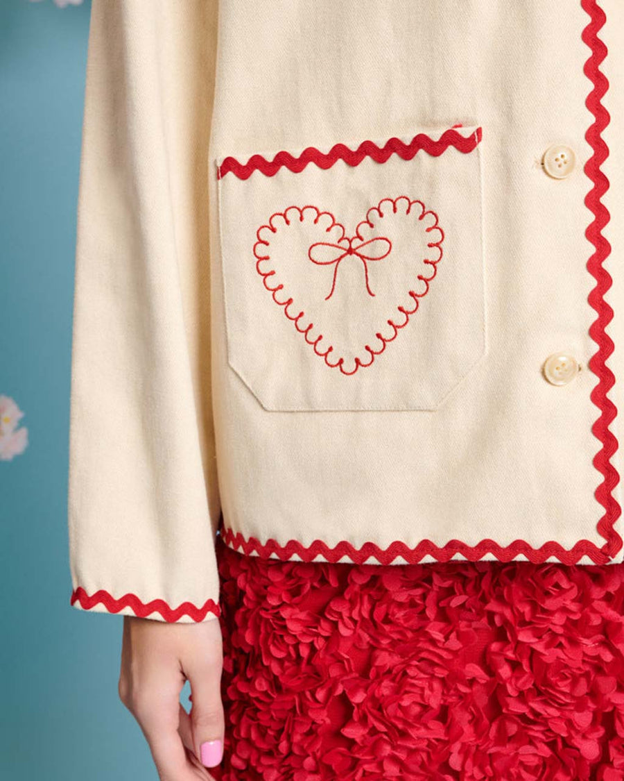 up close of model wearing natural jacket with red ric rac trim and embroidered heart detail on front patch pockets