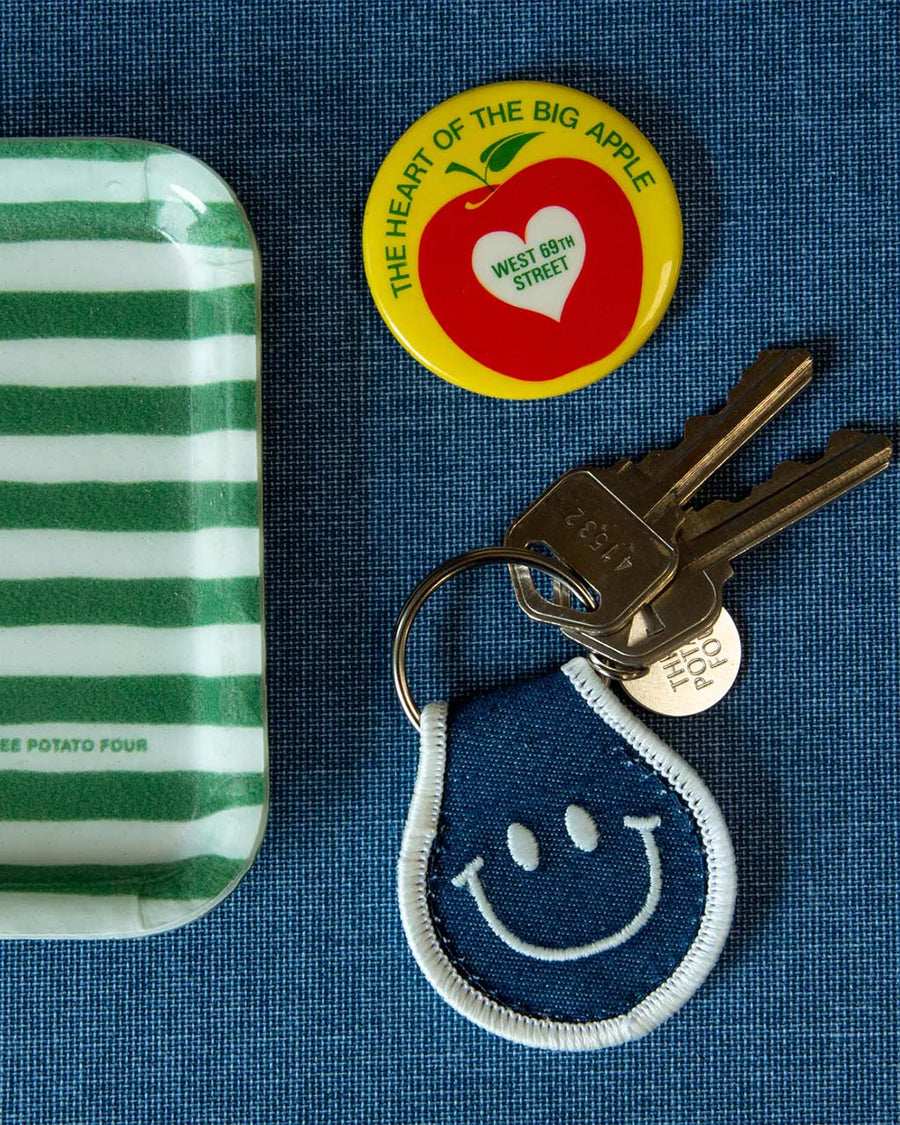denim patch keychain with white smiley face and trim next to NYC pin and striped tray