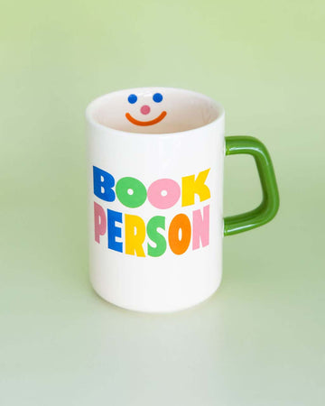 cream ceramic coffee mug with green handle and colorful 'book person' across the front and smiley face inside on green background