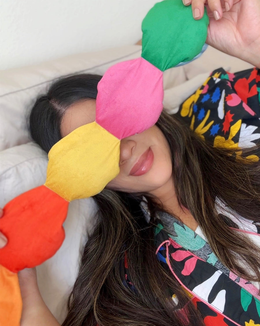 Woman in colorful pajamas holding rainbow colored weighted eye mask towards camera