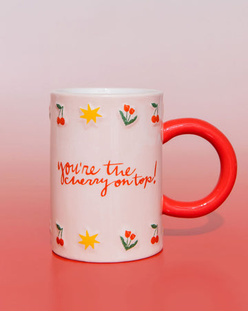 White mug with red handle, with cherry, start and tulip illustrations, that says "you're the cherry on top!" on a red gradiant background