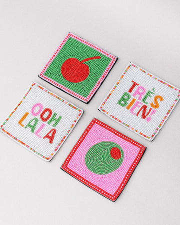sideview of set of 4 beaded coasters: green beaded with cherry print, white with colorful beaded 'tres bien!', white with colorful beaded 'ooh lala', and pink with beaded olive.