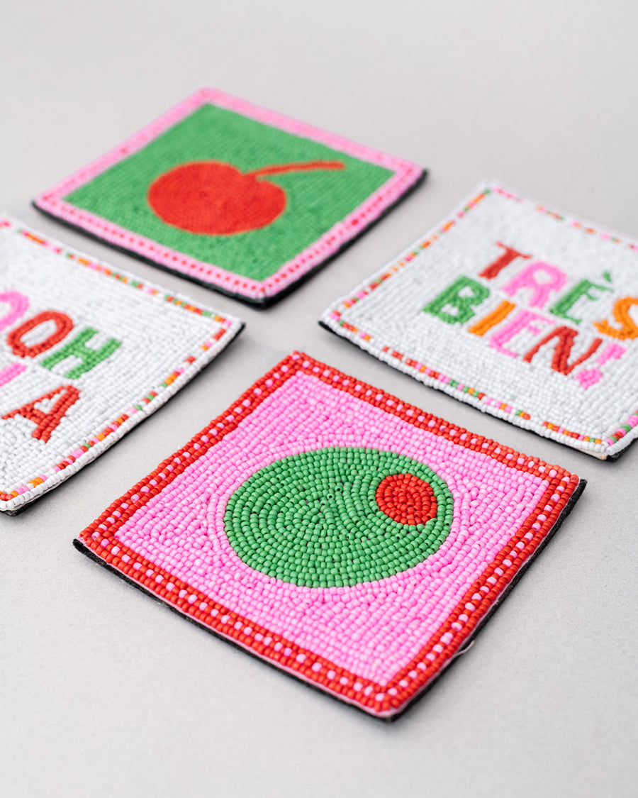 up close of set of 4 beaded coasters: green beaded with cherry print, white with colorful beaded 'tres bien!', white with colorful beaded 'ooh lala', and pink with beaded olive.