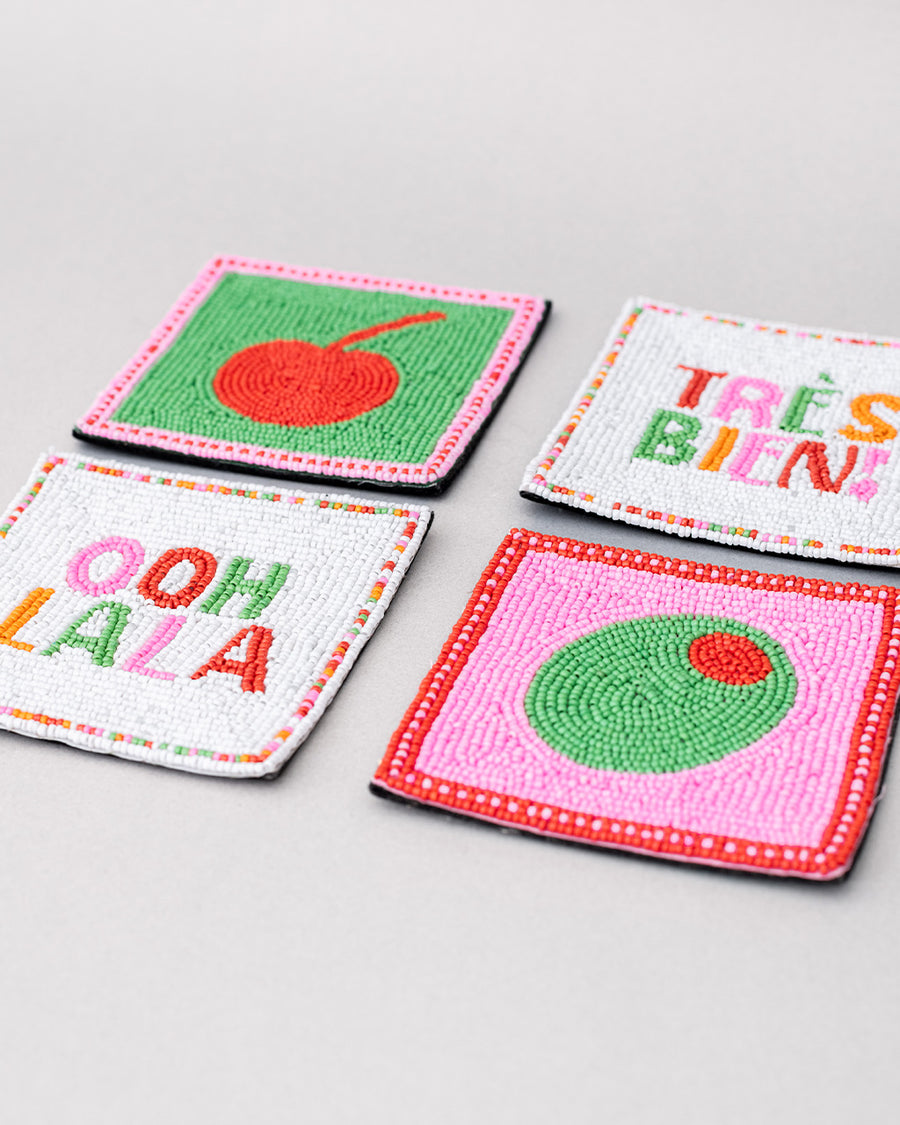 set of 4 beaded coasters: green beaded with cherry print, white with colorful beaded 'tres bien!', white with colorful beaded 'ooh lala', and pink with beaded olive.