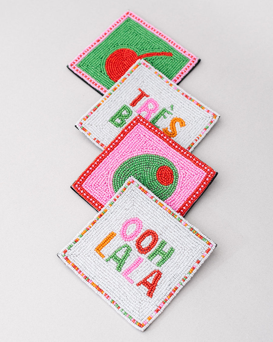 side stacked set of 4 beaded coasters: green beaded with cherry print, white with colorful beaded 'tres bien!', white with colorful beaded 'ooh lala', and pink with beaded olive.