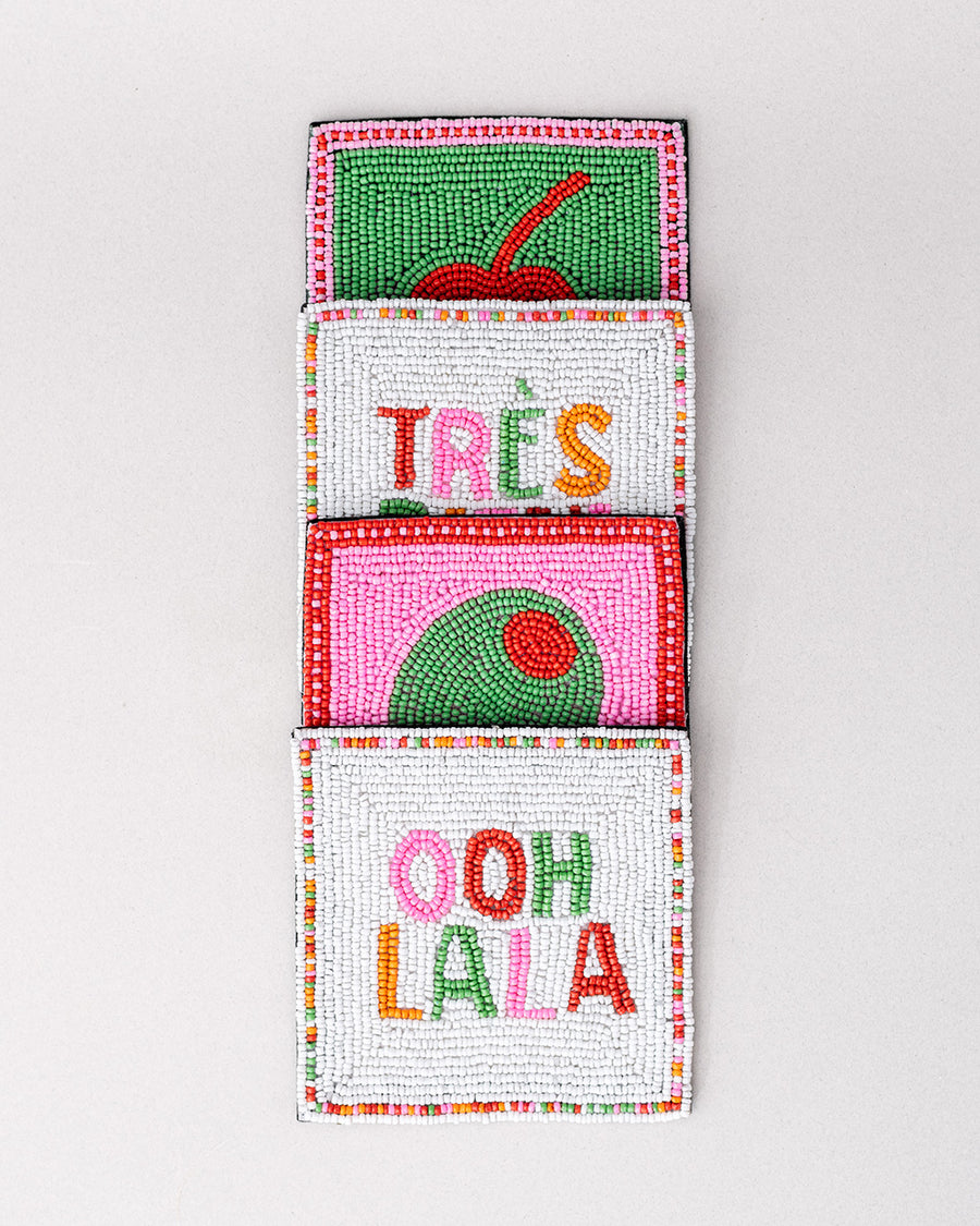 stacked set of 4 beaded coasters: green beaded with cherry print, white with colorful beaded 'tres bien!', white with colorful beaded 'ooh lala', and pink with beaded olive.