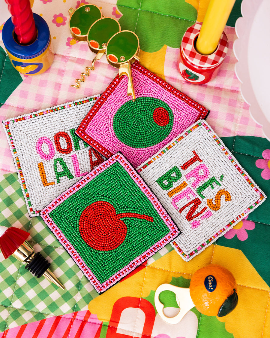 sideview of set of 4 beaded coasters: green beaded with cherry print, white with colorful beaded 'tres bien!', white with colorful beaded 'ooh lala', and pink with beaded olive on a colorful blanket