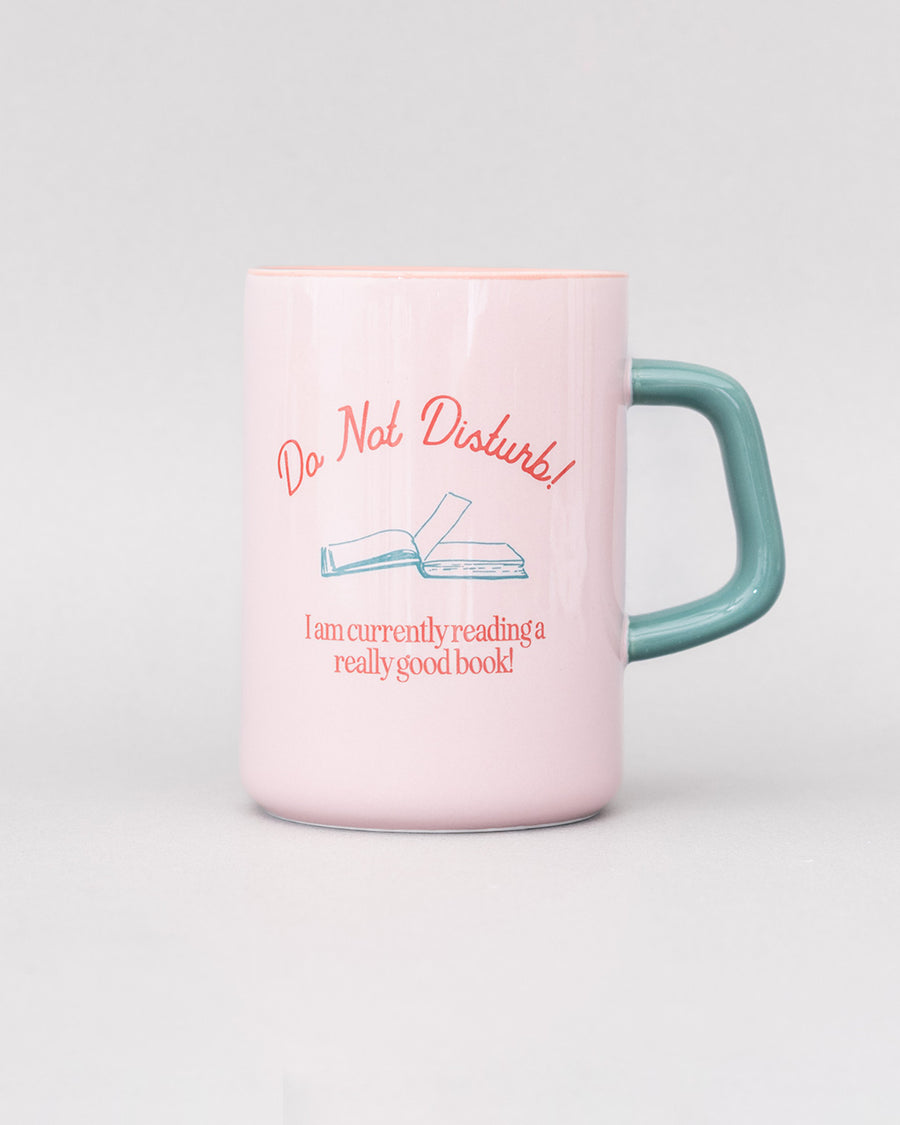 front view of light pink ceramic mug with teal handle and 'do not disturb! i am currently reading a really good book!' typography on both sides