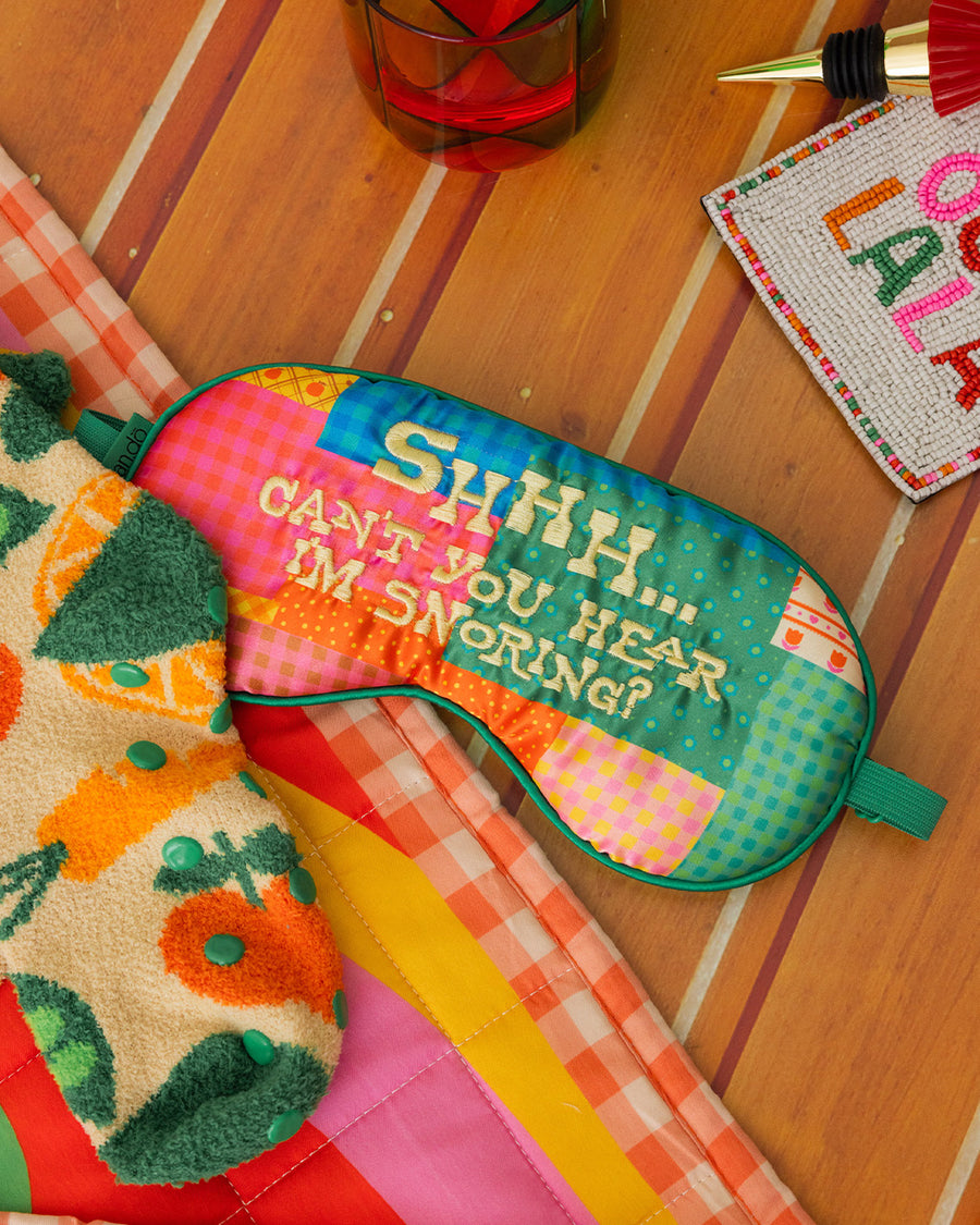 colorful patchwork eye mask with cream 'shhhh...can't you hear i'm snoring?' across the front on a table