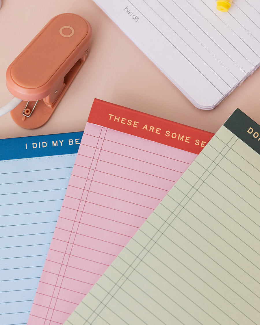 set of three ini legal pads: red/pink with 'these are some serious notes' on the top, green with 'don't forget! write it down!' and blue with 'i did my best' laid flat on a table fanned on a table