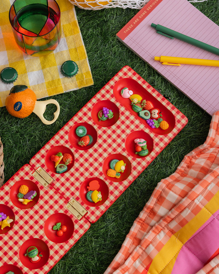 red and cream gingham mancala board with colorful fruit game pieces in grass