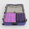set of two packing cubes: medium deep purple with light blue smiley print and large pink with cobalt smiley print in a suitcase