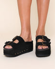 model wearing black suede platform slide sandals with oversized buckles and grommets