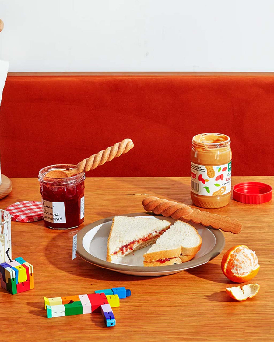 wooden spread spoon and knife with twisted handles on a table with jars of peanut butter and jelly