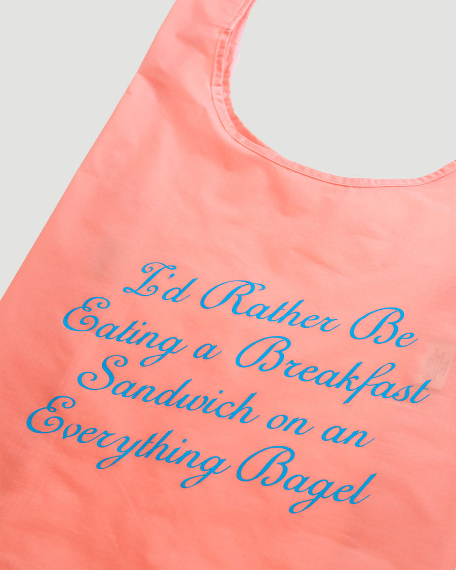 side view of pink standard baggu that says 'i'd rather be eating a breakfast sandwich on an everything bagel' in blue cursive font