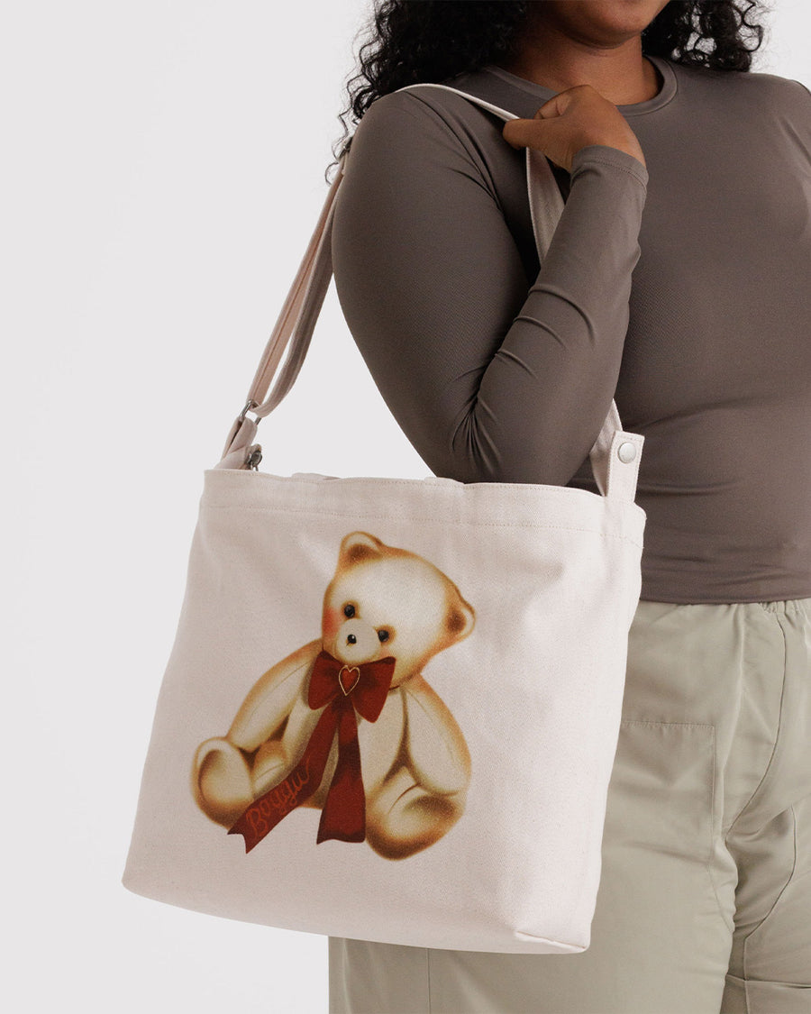 model carrying tan horizontal zip bag with teddy bear with a red bow around the neck