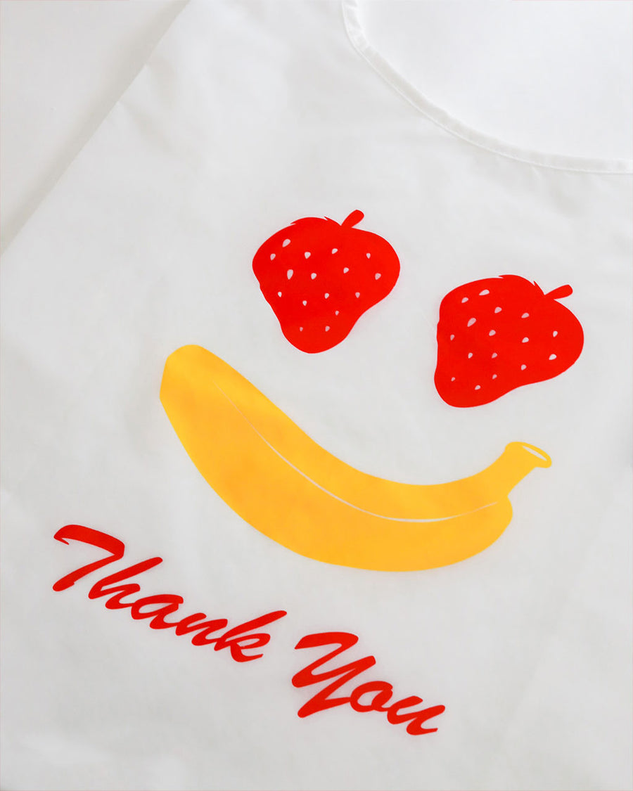 side view of side view of white standard baggu with fruit smiley (strawberry eyes and banana smile) with red 'thank you'