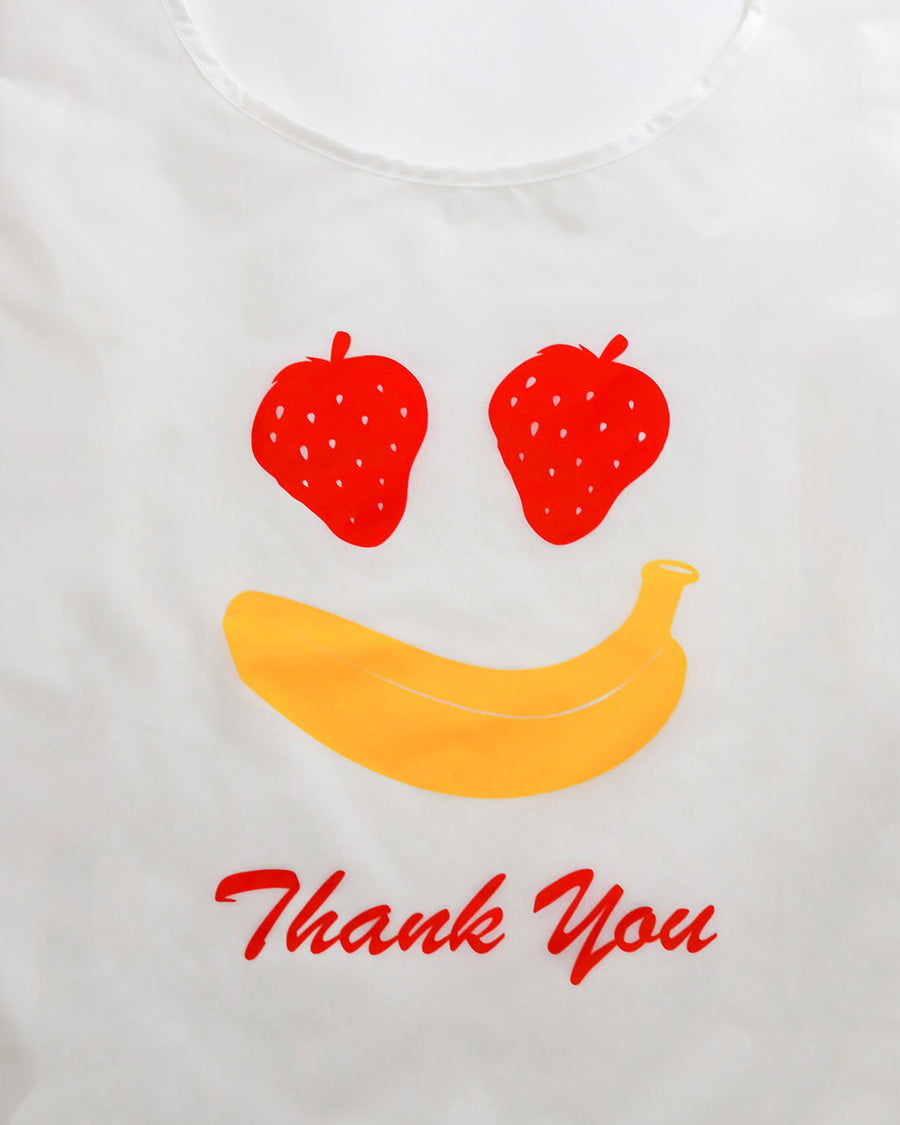 up close of side view of white standard baggu with fruit smiley (strawberry eyes and banana smile) with red 'thank you'