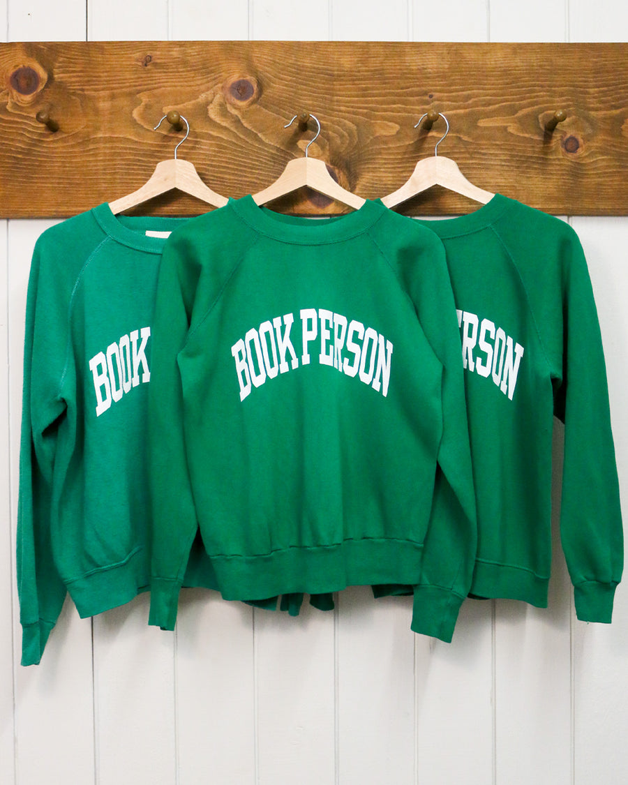 kelly green sweatshirts with white 'BOOK PERSON' across the front hanging on hangers