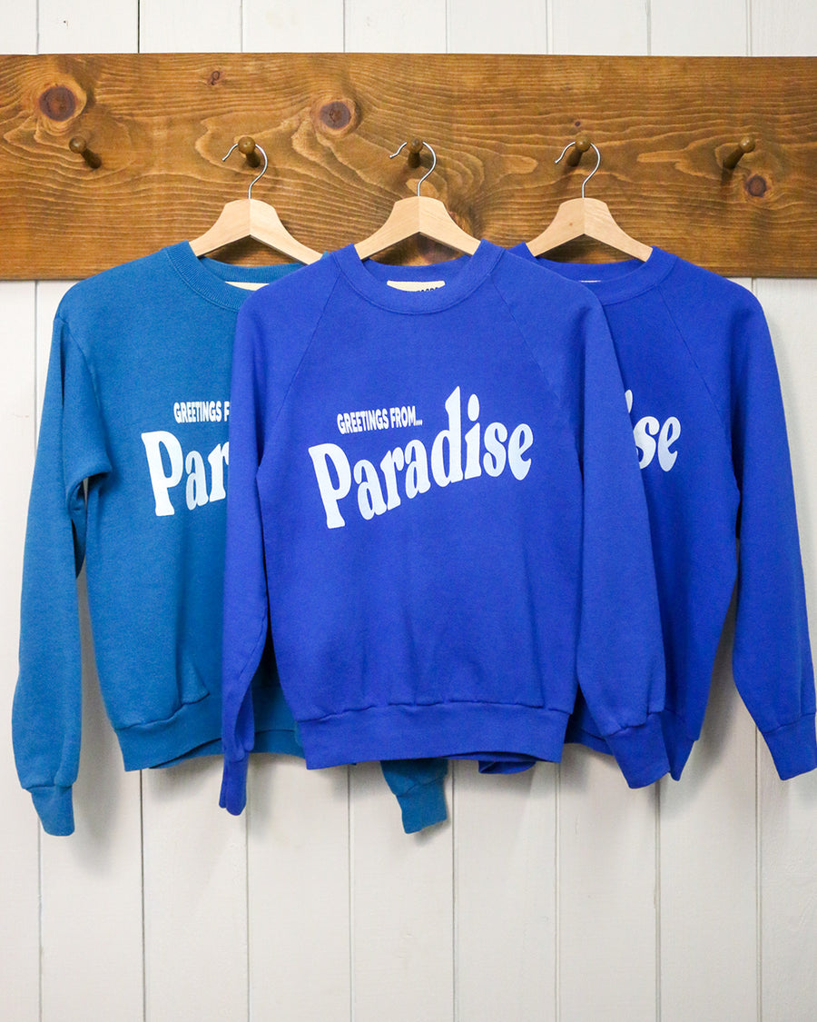 blue sweatshirt with white 'GREETING FROM...PARADISE' across the front hanging on a hanger