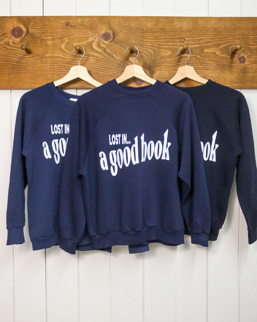 navy sweatshirt with white 'LOST IN... GOOD BOOK' across the front hanging on a hanger