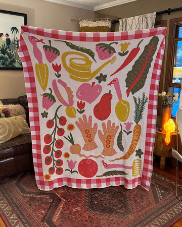 white throw blanket with checkered pink border and various colorful fruits and gardening tools