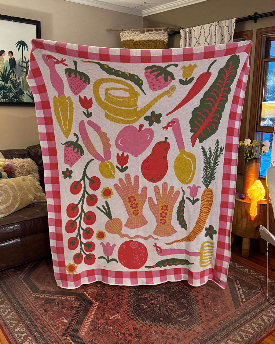 white throw blanket with checkered pink border and various colorful fruits and gardening tools