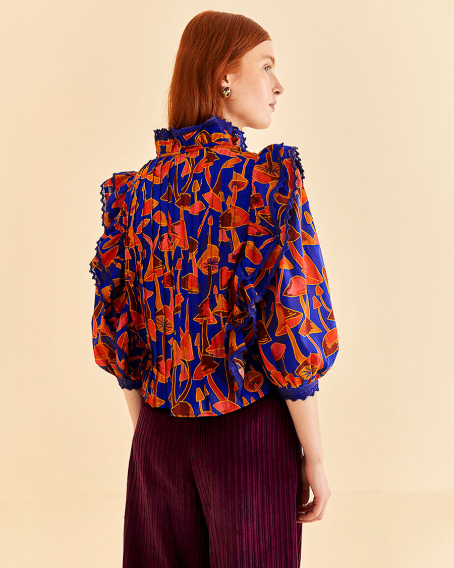 backview of model wearing cobalt blouse with high neck, ruffle sleeves, and all over orange and pink mushroom print