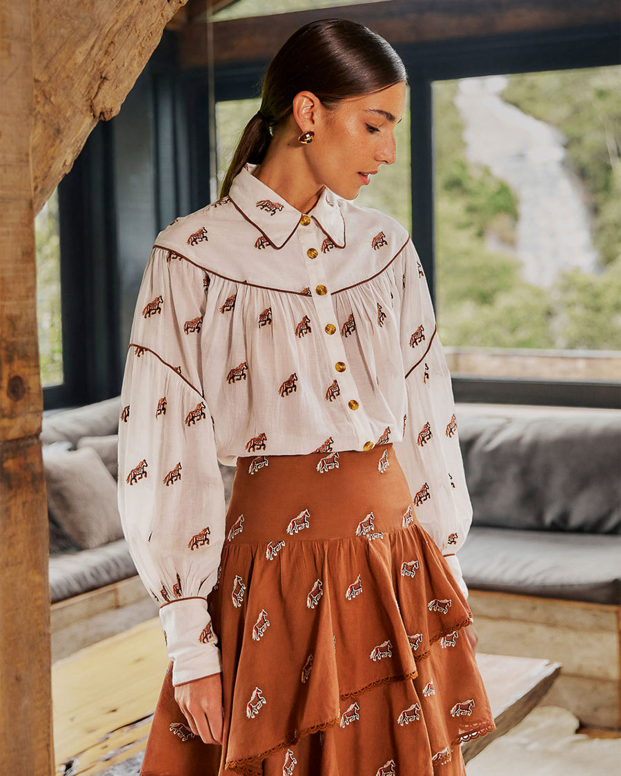 model wearing white button down top with brown piping, tortoise buttons and all over horse print tucked into orange skirt