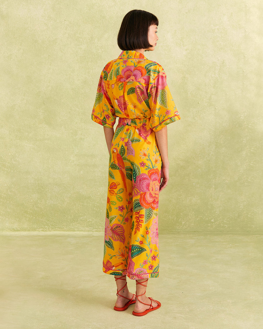 backview of model wearing yellow cropped pants with vibrant flower print, pink piping trim, and pink and yellow flower button and matching top