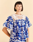model wearing blue and white jungle top with pom pom detail, ruffle sleeves and crochet neckline