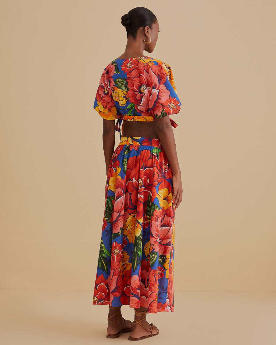 back view of model wearing blue cotton midi skirt with yellow, red and orange cold floral print, side pockets and tie waist and matching cropped top