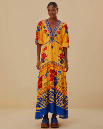 model wearing yellow maxi dress with floral pattern, blue trim details, pom pom detail on the neckline and unique tie front detail