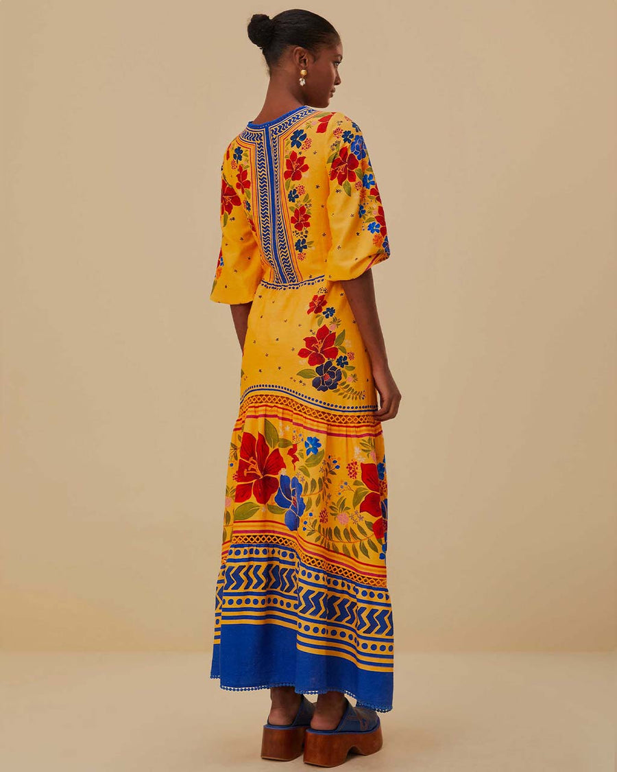 back view of model wearing yellow maxi dress with floral pattern, blue trim details, pom pom detail on the neckline and unique tie front detail
