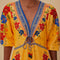 up close of model wearing yellow maxi dress with floral pattern, blue trim details, pom pom detail on the neckline and unique tie front detail