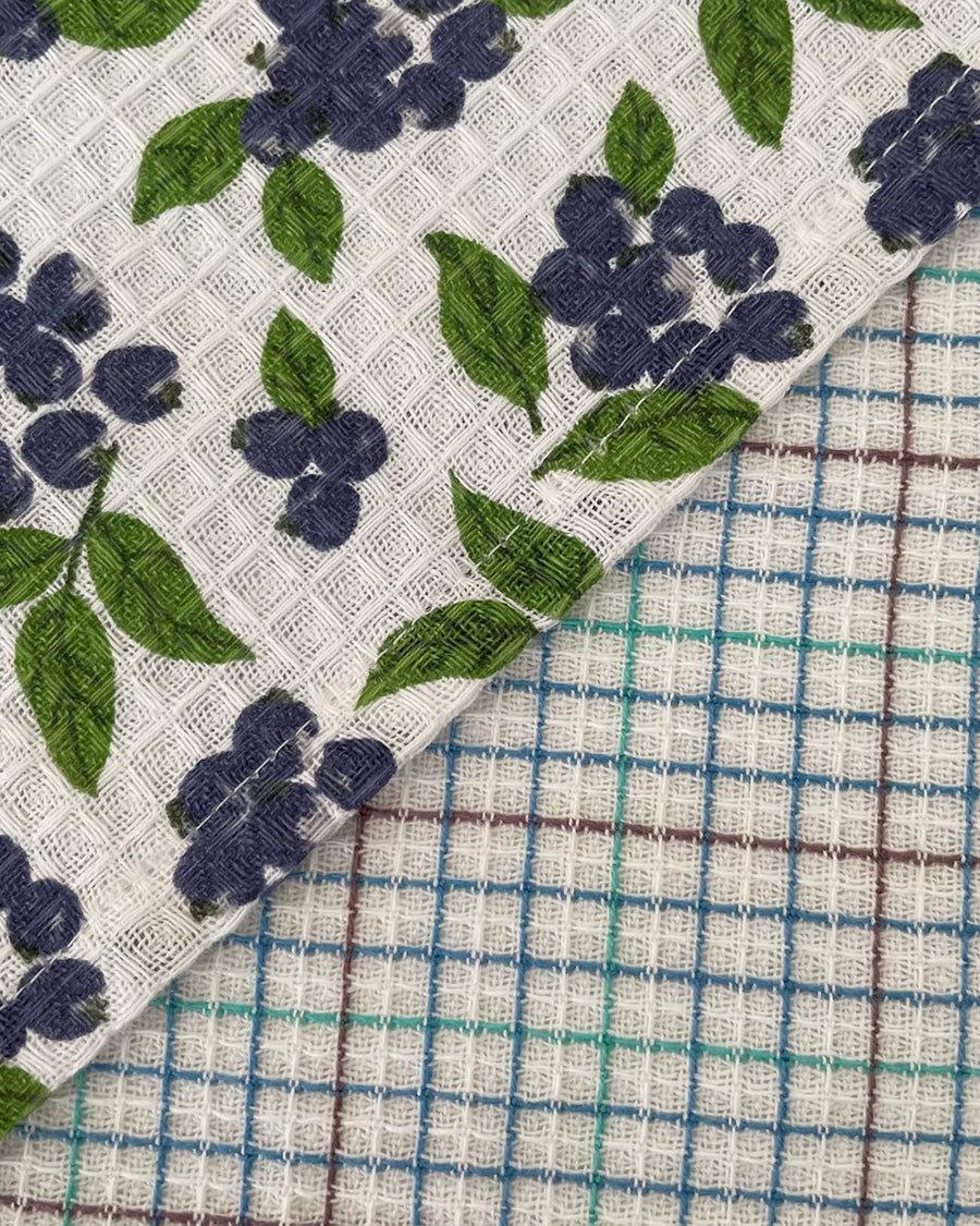 up close of set of two woven dishcloths: white with blueberry print and white with plaid in blue, green and burgundy