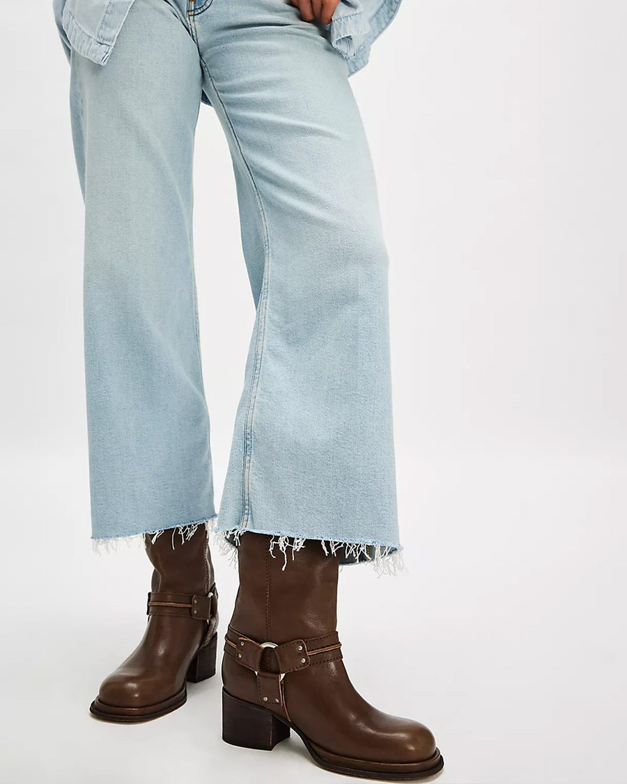 model wearing tall dark brown leather boots with silver buckle and strap detail around the ankle with light denim frayed jeans