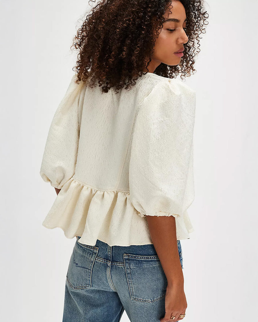 back view of model wearing jacquard blouse with puff sleeves, slight peplum hem, and black bow and buttons on the bust