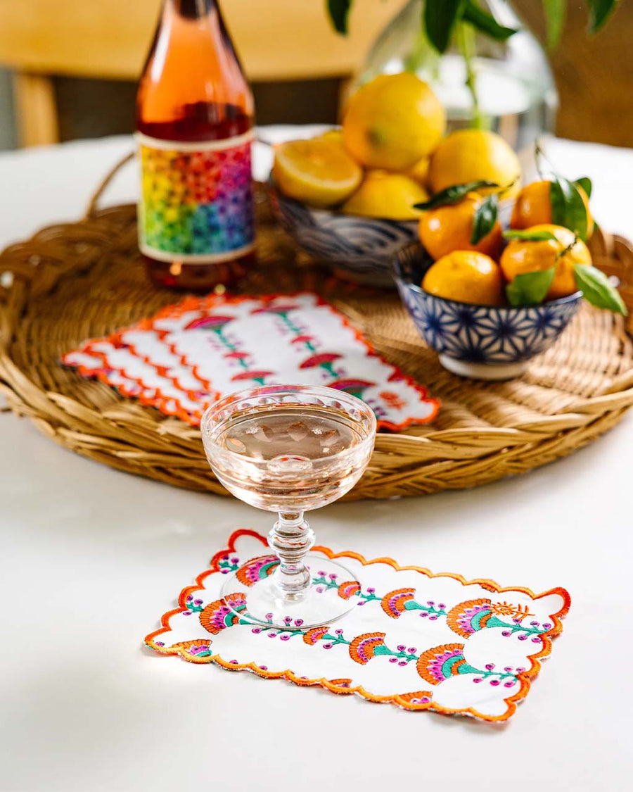 colorful print cocktail napkin with orange embroidered scalloped edges with a glass on it