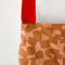up close of light tan wine bag with terracotta abstract checkers and red woven strap