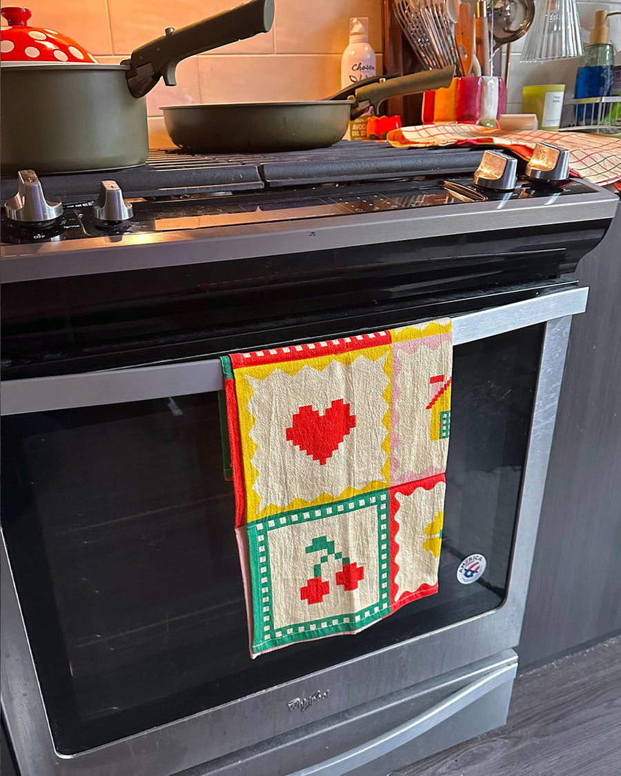 cream tea towel with strawberry, shamrock, banana, heart, house, pretzel, cherry, flower and bow icons hanging on an oven handle