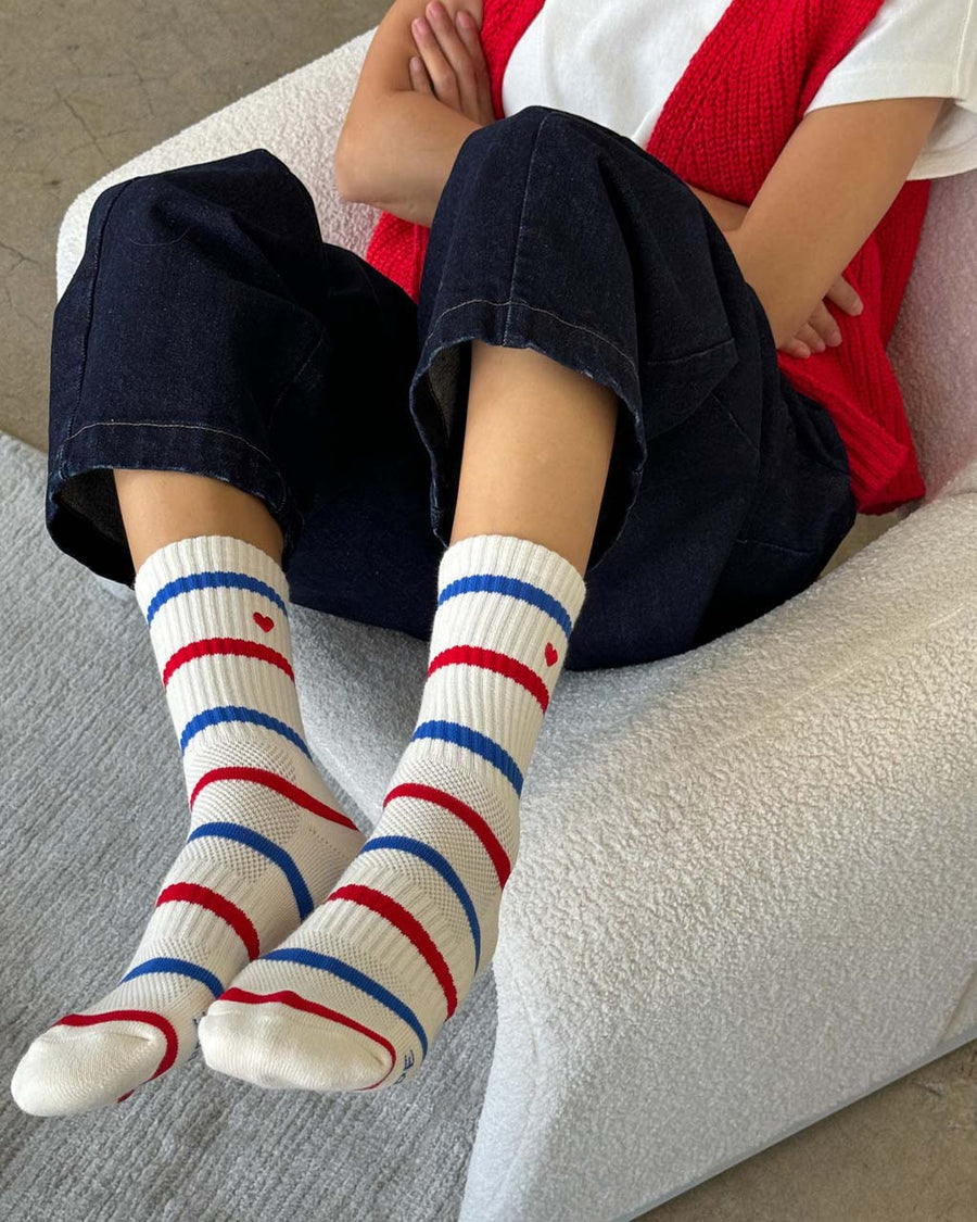 model wearing white socks with red and blue multiple stripe socks with embroidered red heart on a chair